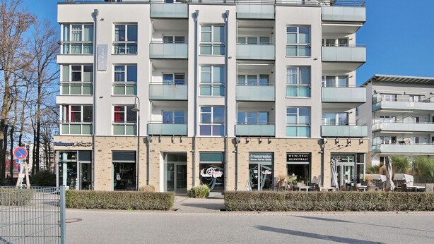 Un hotel de varios pisos con diseño moderno, con balcones en cada planta, un restaurante a nivel de calle y un cartel visible de un establecimiento cercano.