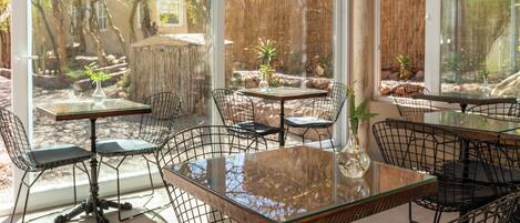 An outdoor dining area with glass tables and wire mesh chairs, surrounded by a bamboo fence and various plants.