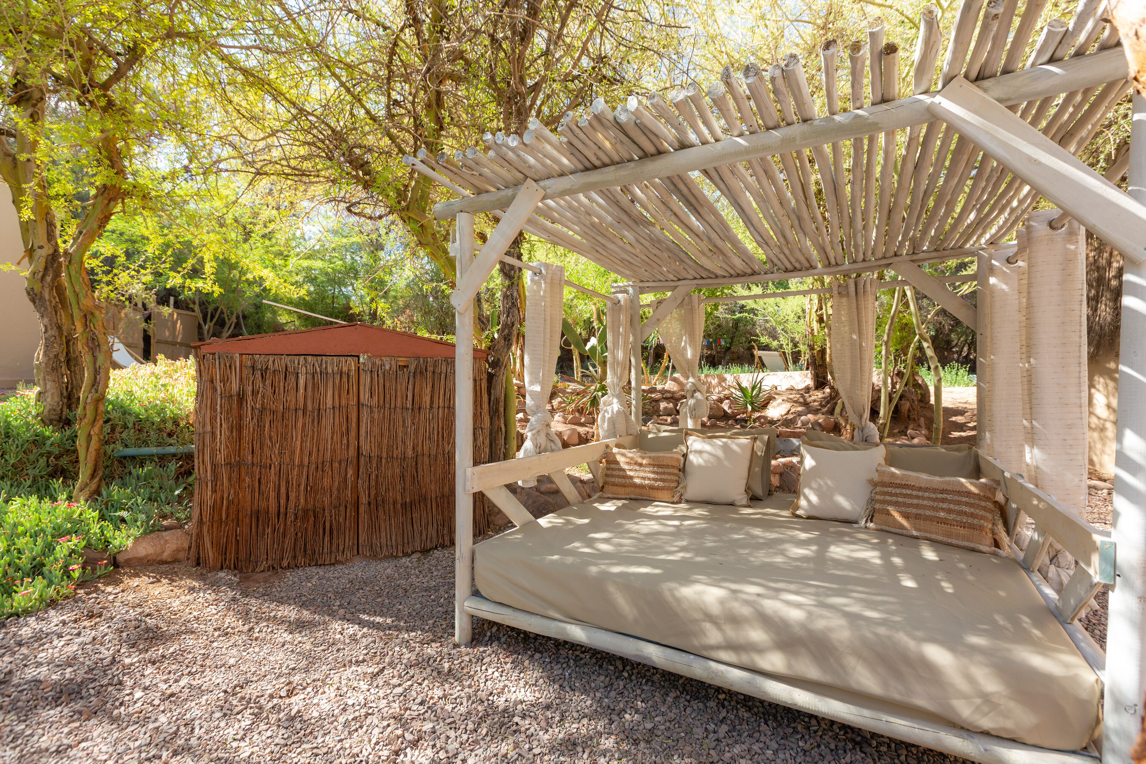 A covered outdoor seating area with a bench and cushions, surrounded by trees and a thatched structure.