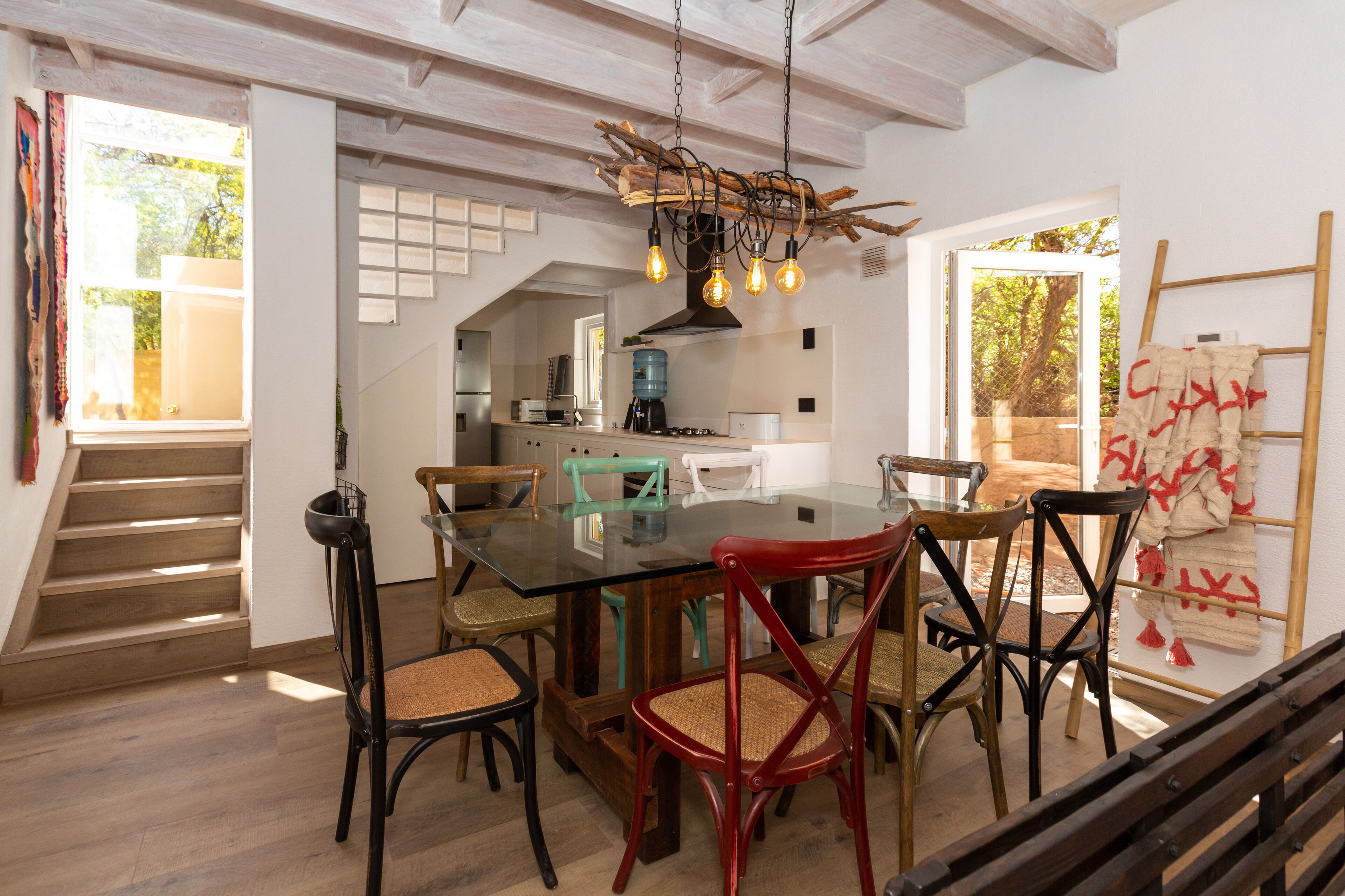 A dining area with a glass table and chairs, a hanging light fixture, and a staircase leading to another room.
