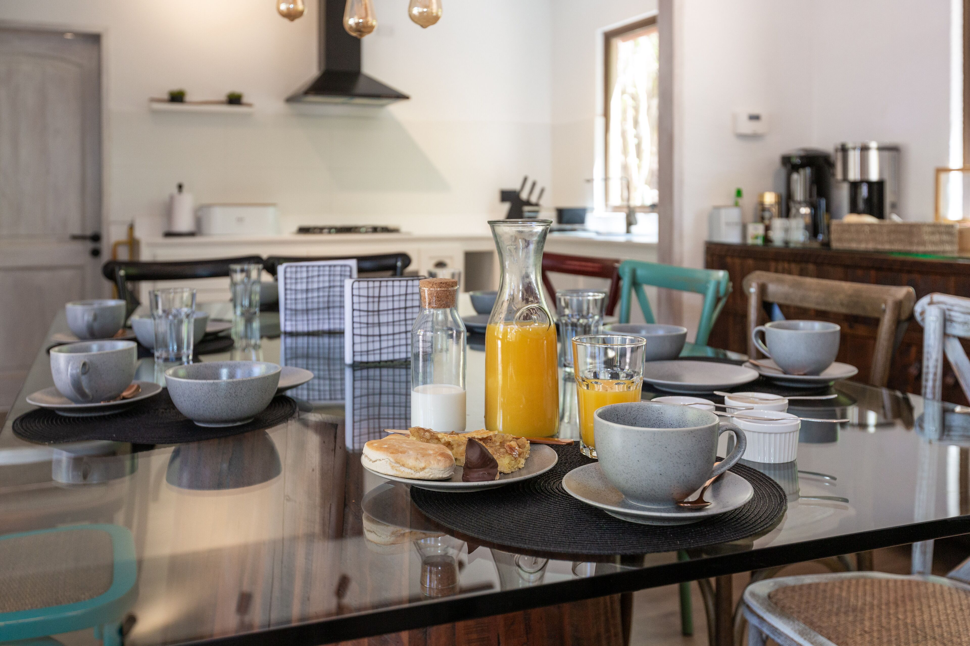 Une table mise pour le petit-déjeuner, avec une table en verre, des assiettes, des tasses et un pichet de jus d’orange.