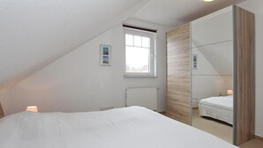 Un dormitorio con techo inclinado, un ventanal grande, un armario con espejo y una cama con edredón blanco.