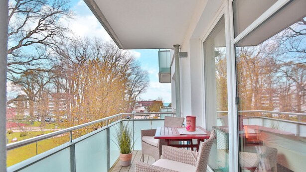 Un balcon agrémenté de meubles en osier, d’une plante en pot et d’un récipient rouge.