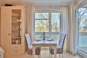 A dining area with a table set for two, a cabinet with glass doors, and a view of trees through large windows.