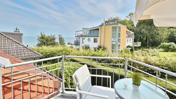 A balcony with a glass table and white chair, overlooking a residential area with a view of the sea.