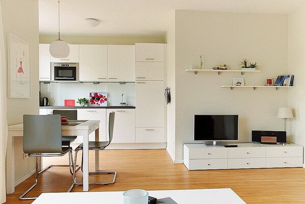 A modern kitchen with white cabinets, a dining area with a table and chairs, a TV on a stand, and shelves with decorative items.