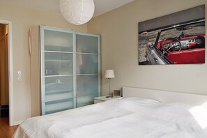 A bedroom with a bed, a nightstand, a lamp, a wardrobe, and a wall-mounted painting of a car.