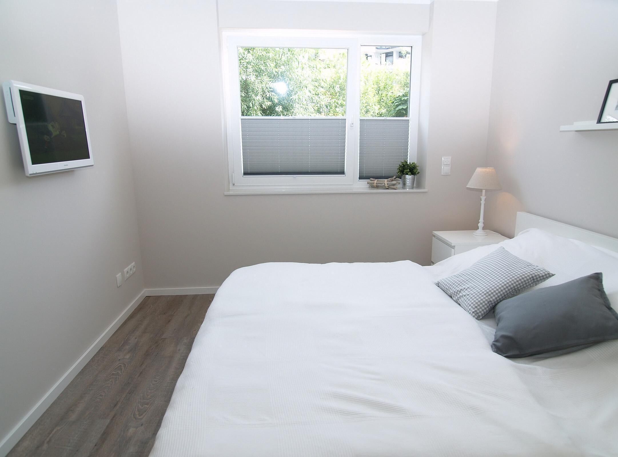 A bedroom with a bed, a bedside table with a lamp, a window with blinds, and a wall-mounted TV.