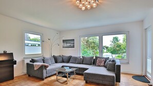 A modern living room with a sectional sofa, a coffee table, and a fireplace. There are large windows providing natural light and views of the outdoors. - Ferienhaus Ragna Appartement 1 (Haffkrug)