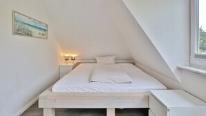 Una camera da letto con soffitto spiovente, un letto con lenzuola bianche, un comodino e un quadro appeso al muro.