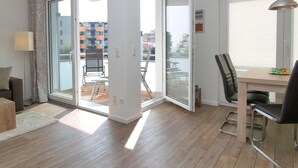 Un apartament modern amb terres de fusta, zona de menjador amb taula i cadires, sofà i una porta corredissa de vidre que dona a un balcó amb mobles d'exterior.