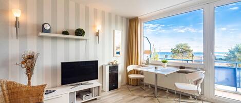 A modern living room with a TV, sofa, dining area, and large windows offering a view of the sea.
