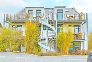 Ein modernes, zweistöckiges Haus mit Wendeltreppe, Balkonen mit Gartenmöbeln und einem geparkten Auto davor.