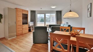 Una sala de estar moderna con piso de madera, sofás grises, una mesa de comedor de madera y un ventanal con cortinas.