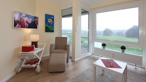 Una sala de estar moderna con un ventanal grande, un sillón cómodo, una mesita pequeña y un televisor en la pared.