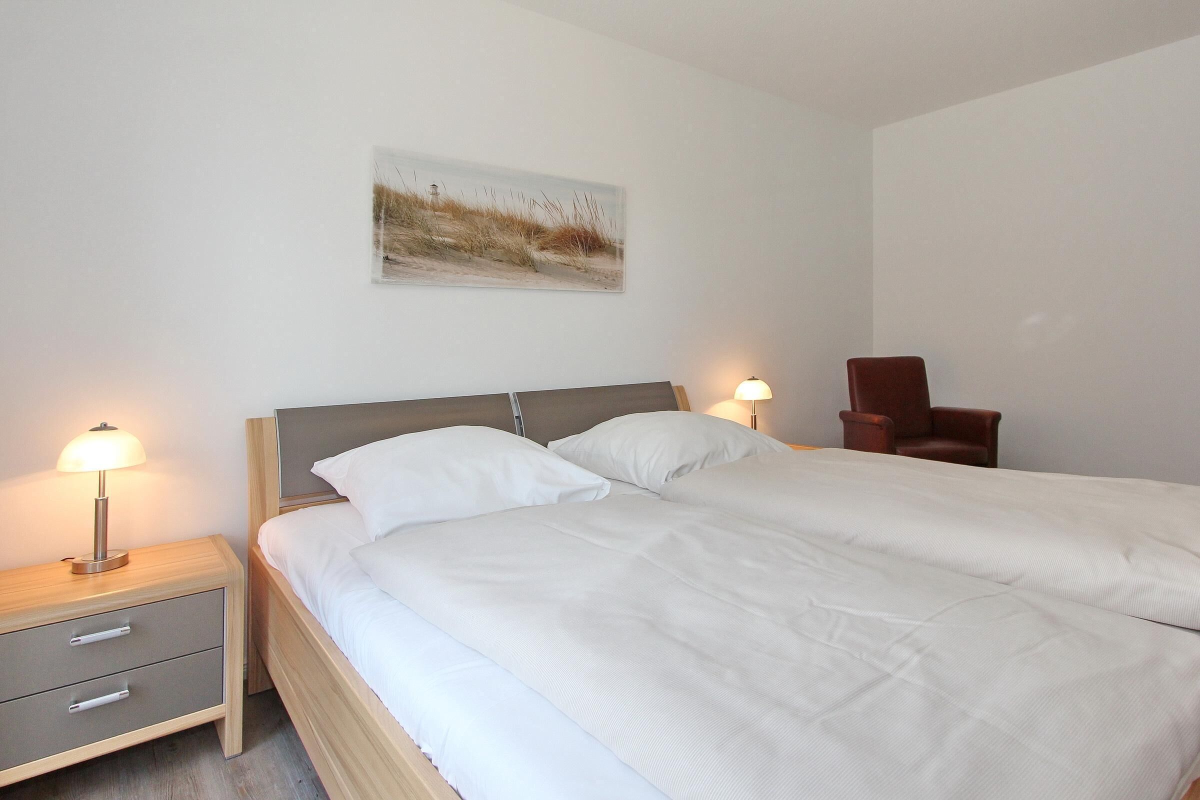 A neatly made bed with white linens and a wooden headboard, a nightstand with a lamp, and a brown chair in a simple room with a single wall-mounted picture.