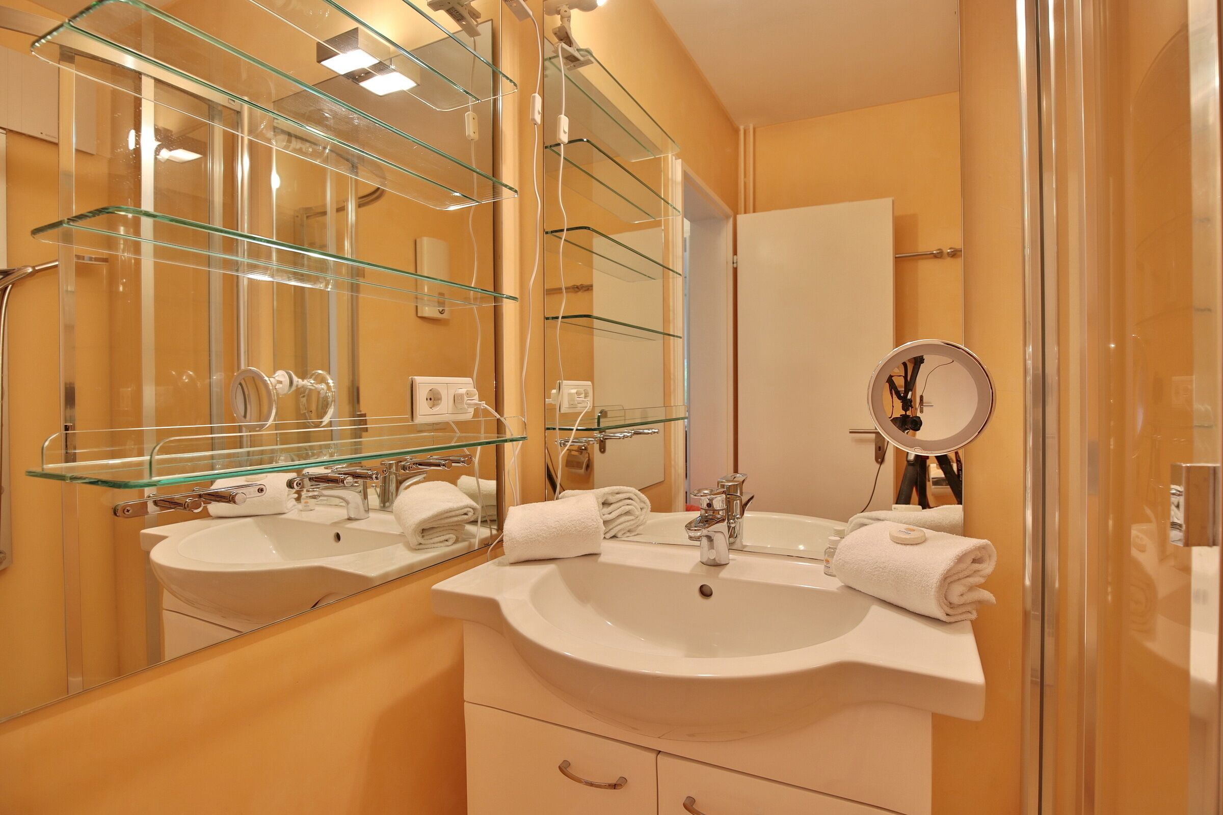 Een badkamer met twee wastafels, glazen planken, een grote spiegel en een douchecabine van glas.