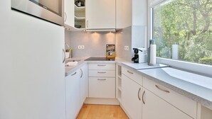 A modern kitchen with white cabinets, a countertop, and a window with a view of trees. - Strand und Meer Appartement 1 (Scharbeutz)