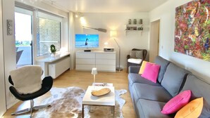 A modern living room with a grey sofa, a white coffee table, a black chair, a cowhide rug, a television, and a colorful abstract painting. - Strand und Meer Appartement 1 (Scharbeutz)