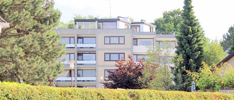 A multi-story residential building with balconies, surrounded by well-maintained hedges and trees.