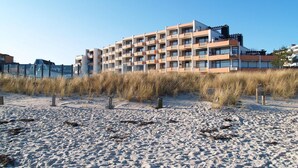 Un hôtel à plusieurs étages, doté de balcons, situé juste à côté d’une plage de sable bordée d’herbes hautes.