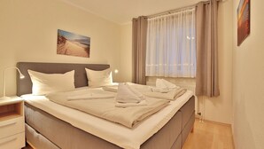 Een netjes opgemaakt bed met kussens en dekbed, een nachtkastje met een lamp, een raam met gordijnen, en een schilderij aan de muur.