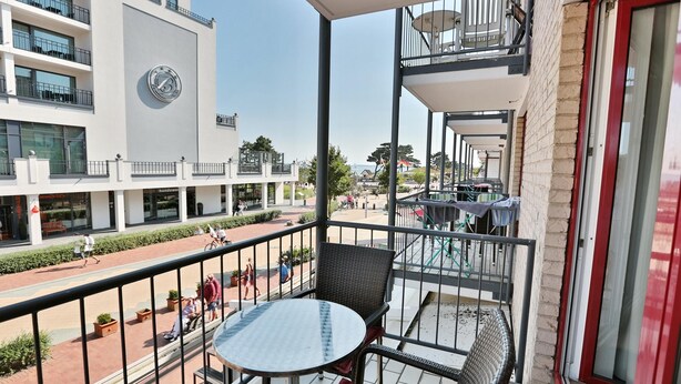Een balkon met een ronde tafel en rieten stoelen, met uitzicht op een voetgangersstraat met gebouwen en een heldere lucht.
