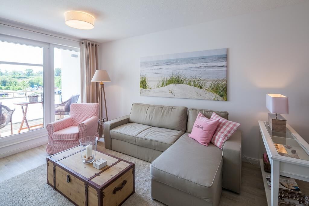 En hyggelig stue med sofa, lænestol og et lille bord med lampe. Et maleri af en strand hænger på væggen, og der er udsigt til grønne områder udenfor.