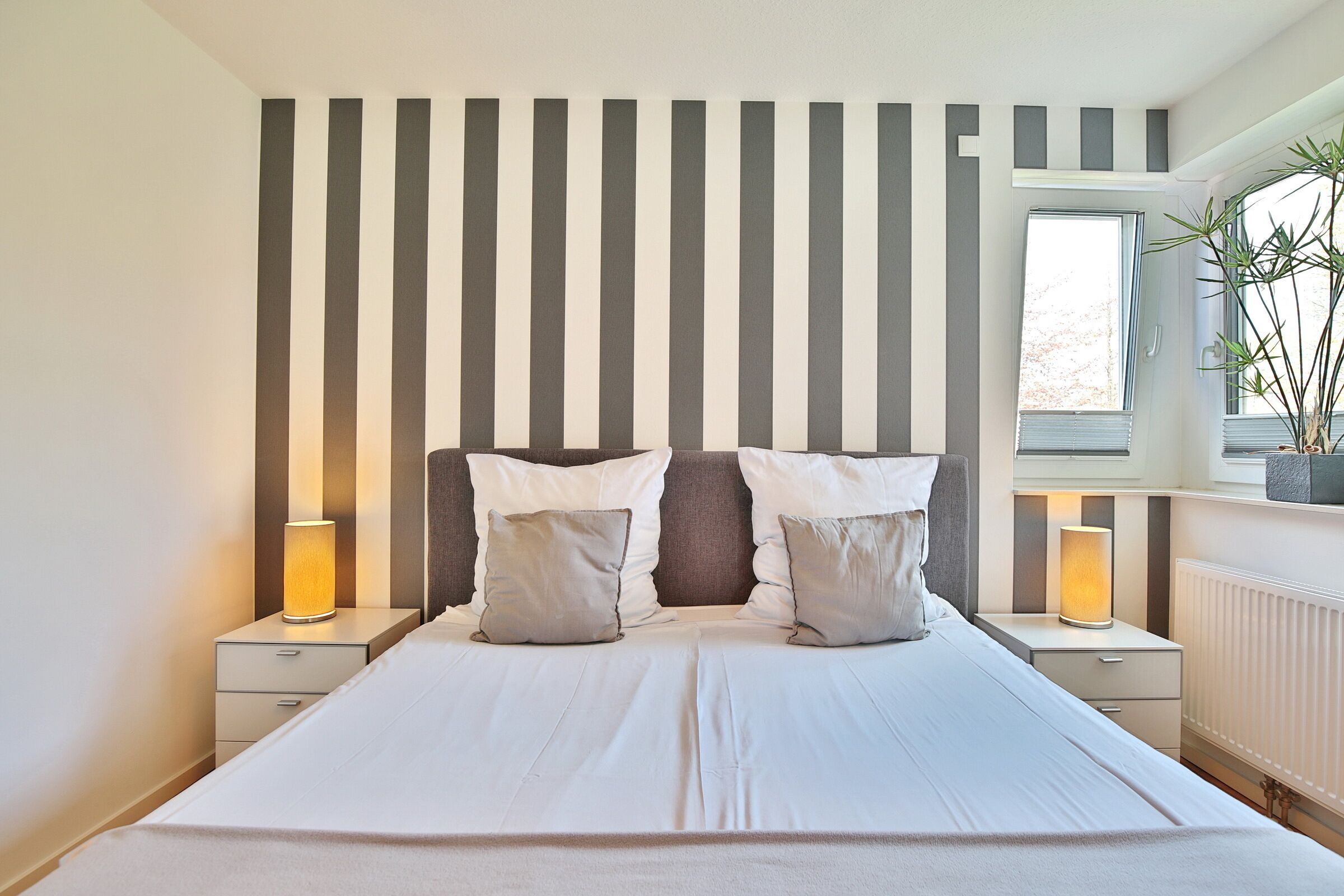 A bedroom with a large bed, two bedside tables, a window with a plant, and striped wall panels.