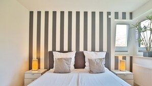 Une chambre avec un grand lit, deux tables de chevet, une fenêtre avec une plante et des panneaux muraux à rayures.