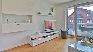 Un salon moderne doté d’un téléviseur à écran plat, de meubles blancs, d’une table basse en verre et offrant une vue sur un immeuble en briques rouges, visible à travers une grande fenêtre.
