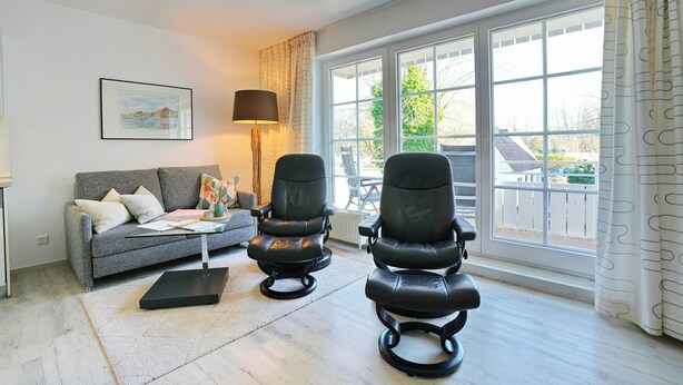 A living room with a grey sofa, two black chairs, a floor lamp, and a large window with a view of a house and trees. - Parkblick Appartement 4 (Scharbeutz)