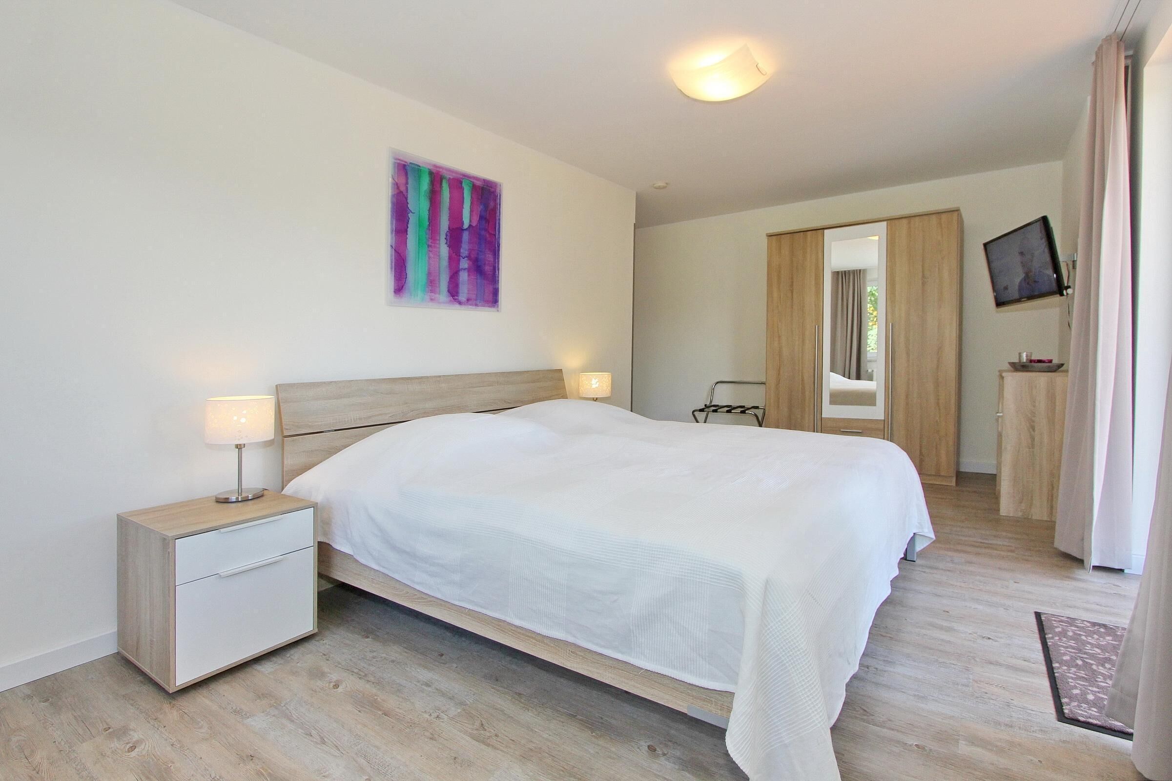 A bedroom with a bed, bedside table, lamp, artwork, TV, and a wardrobe.