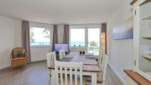 Un comedor con una mesa de madera y sillas blancas, un televisor y vistas al mar a través de amplios ventanales.