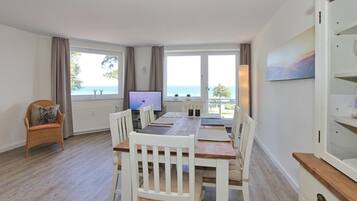 A dining area with a wooden table and white chairs, a television, and a view of the sea through large windows.