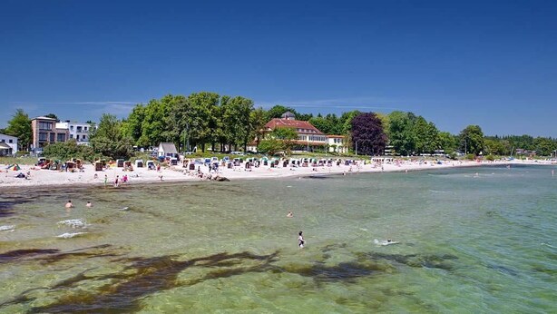 Una playa con aguas cristalinas, gente nadando y edificios al fondo.