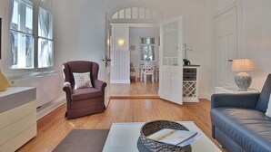 A living room with a brown armchair, a blue sofa, a white coffee table, and a basket with papers. There is a window with curtains, a door leading to another room, and a lamp on a side table. - Villa Schöneck Appartement 1 (Scharbeutz)