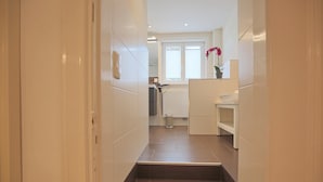 A narrow hallway with a dark brown tiled floor leading to a kitchen area with white cabinets and a window. - Villa Schöneck Appartement 1 (Scharbeutz)