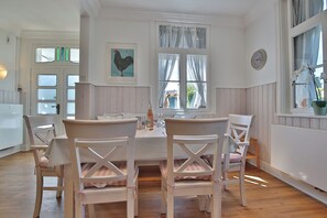 A dining room with a white table and chairs, a window with curtains, and a wall clock. - Villa Schöneck Appartement 1 (Scharbeutz)