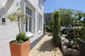 A narrow pathway leading to a white building with windows and a door. There are potted plants along the path and a car parked on the street.
