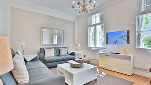 A modern living room with a grey sofa, a white coffee table, a television, and a large window with curtains. - Villa Schöneck Appartement 1 (Scharbeutz)
