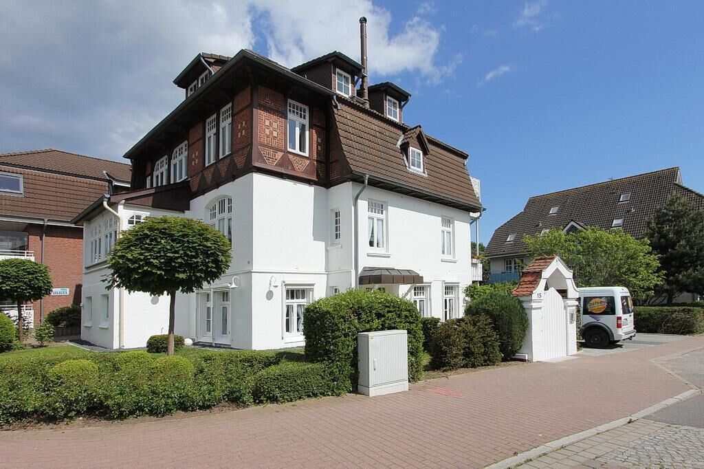 A two-story house with a white facade and brown roof, surrounded by hedges and a paved driveway.