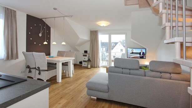 A modern living room with a grey sofa, a dining table, and a TV in the background. - Domizil am Möwenberg Appartement 9 (Scharbeutz)