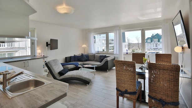 Una sala de estar moderna con un sofá, mesa de comedor y sillas. Cuenta con una zona de cocina con fregadero y armarios. Amplias ventanas ofrecen luz natural y vistas al exterior.