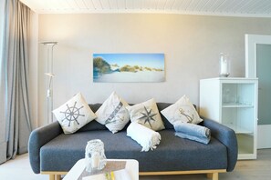 A living room with a grey sofa, nautical-themed cushions, a painting of a beach, a floor lamp, and a glass cabinet. - Seetempel Appartement 27 (Scharbeutz)