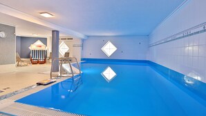Une piscine intérieure à l’eau claire, avec un escalier et un espace ensoleillé aménagé avec des chaises et une table.