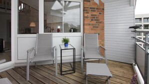 Un balcon doté d’un plancher en bois, de murs blancs et d’un mur d’accent en briques. On y trouve deux chaises en métal et une petite table sur laquelle est posée une plante en pot.