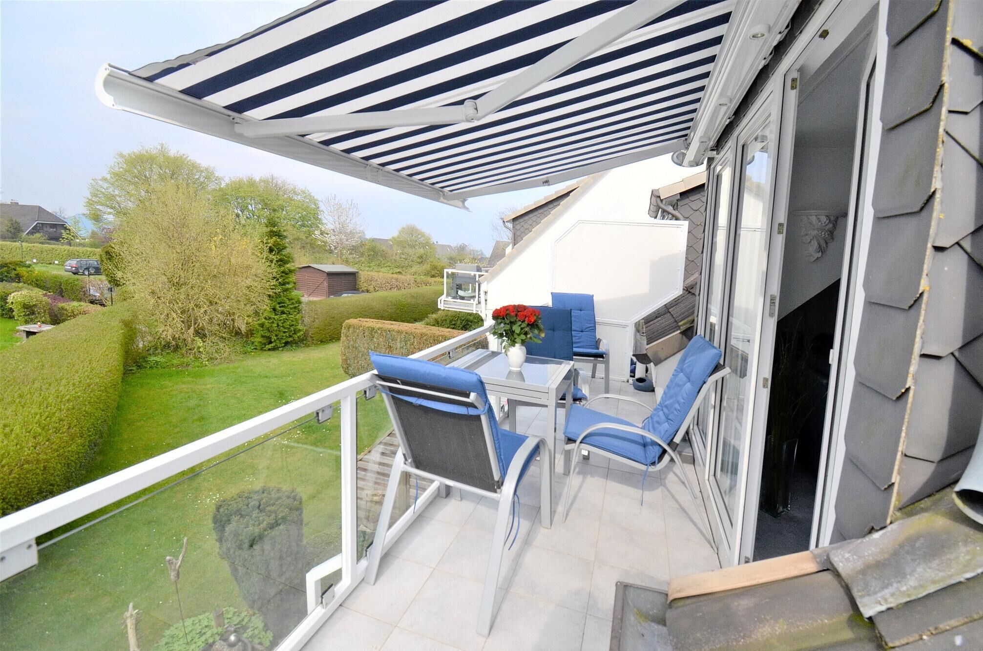 Een balkon met een gestreepte zonnescherm, een glazen tafel, blauwe stoelen en een vaas met rode bloemen.