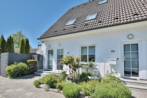 A modern two-story house with a tiled roof, a front door, and a small garden area with various plants and shrubs. - Maisonette mit Charme (Scharbeutz)