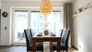 Uma área de refeições com uma mesa posta para quatro pessoas, uma luminária pendente de vime e vista através de uma grande janela.